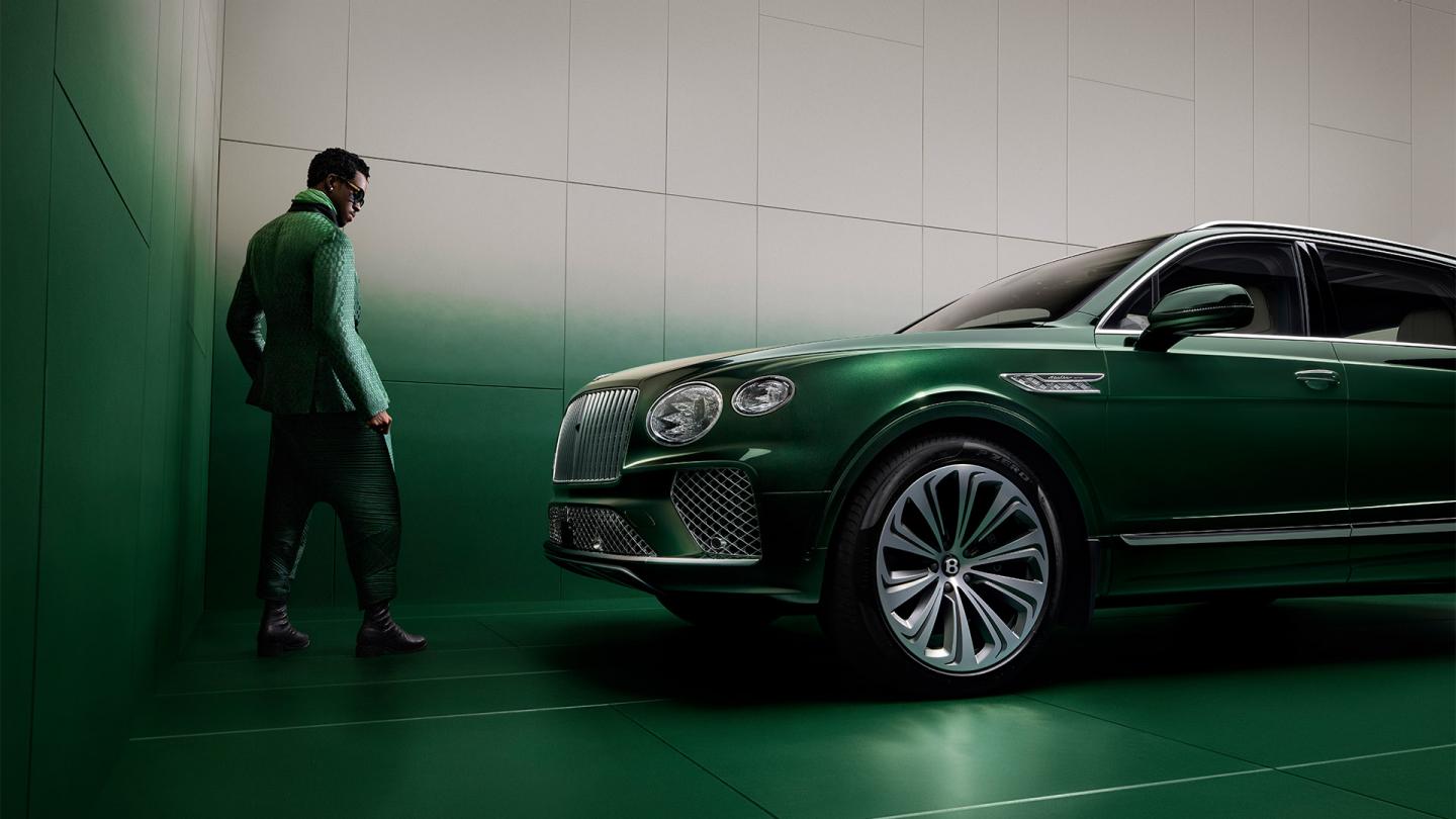 Wisdom Kaye standing next to the front three quarter of the Bentayga Atelier Edition in an Emerald exterior colours with bright chrome accents