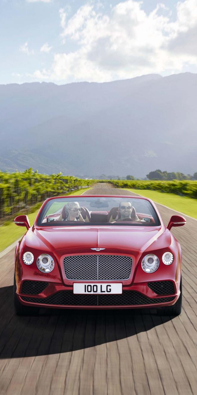 Front side view of Bentley Continental GTC with two passengers in view, driving along green road.
