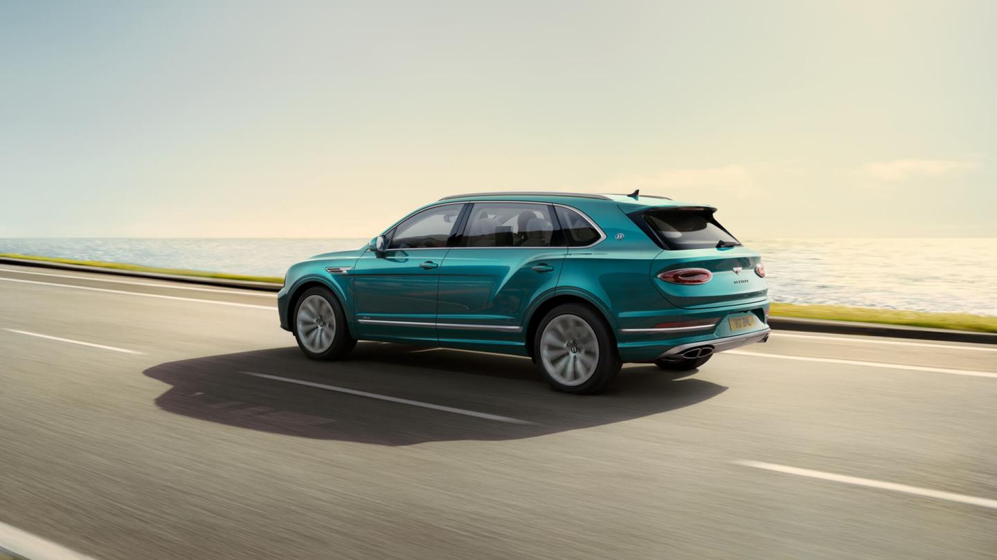 Rear side angled view of Bentley Bentayga EWB Azure in Topaz Blue with chrome accent, driving along sea side.