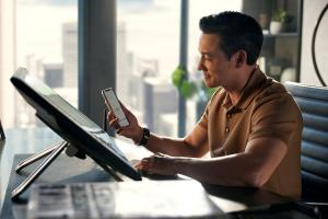A person infront of deskop screen looking at phone displaying My Bentley App.